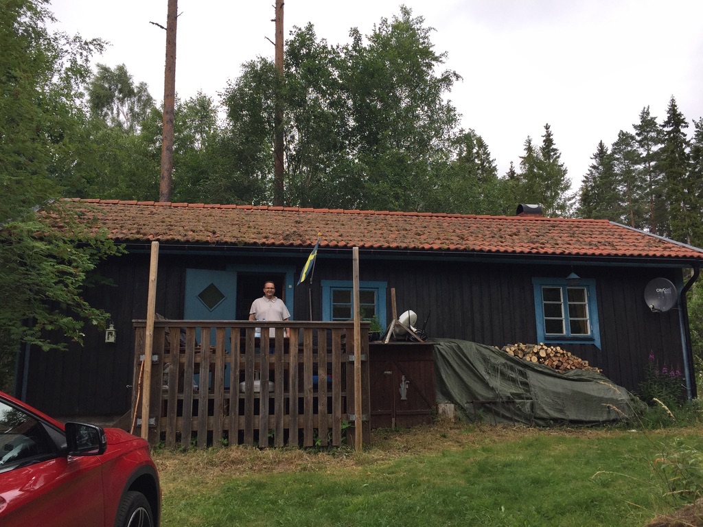 Pilstugan: ein dunkles Holzhaus mit blauen Fensterrahmen und rotem Ziegeldach, auf dem Hof weht die schwedische Flagge, dahinter Wald, davor eine Wiese